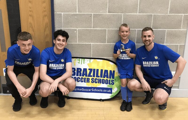 Children's Football. Image shows a group of Brazilian Soccer Schools coaches with a student in front of the BSS sign.