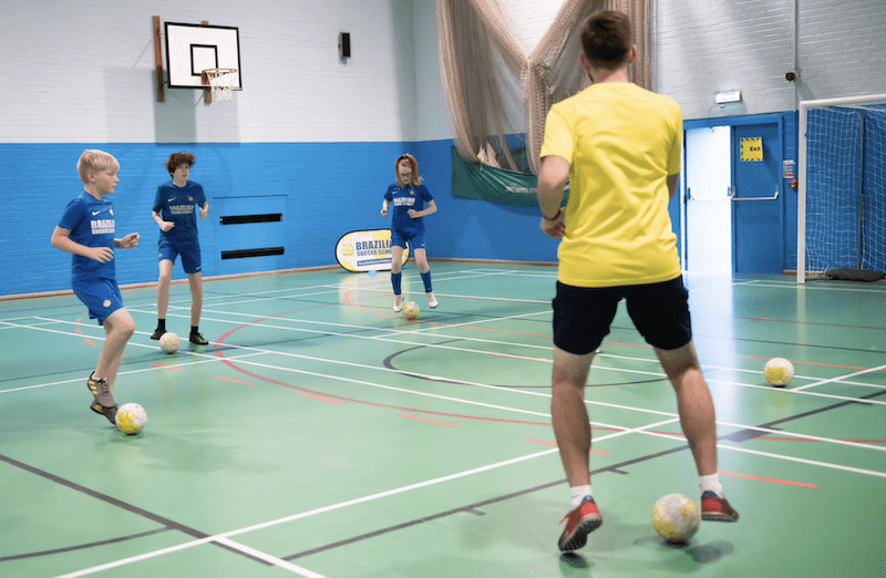 Football training for kids. Image shows 3 children doing drills with a football and a Brazilian Soccer Schools coach. 